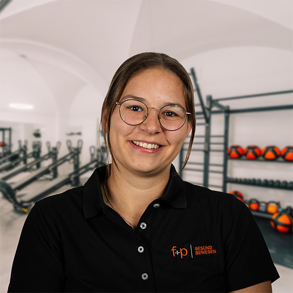 CoralieSchie Junge Frau mit Brille und schwarzem Polo-Shirt in einem Fitnessstudio mit Trainingsgeräten im Hintergrund.