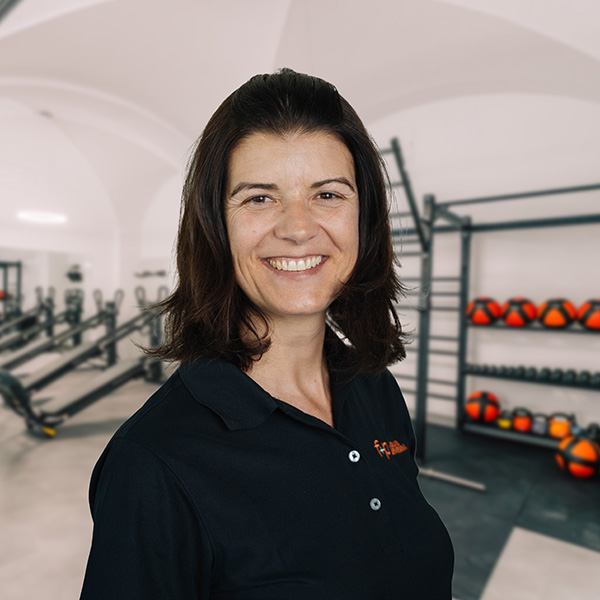 HeiKo Lächelnde Heidrun im schwarzen Poloshirt vor Trainingsgeräten im Fitnessbereich von f+p Gesund Bewegen in Kempten.