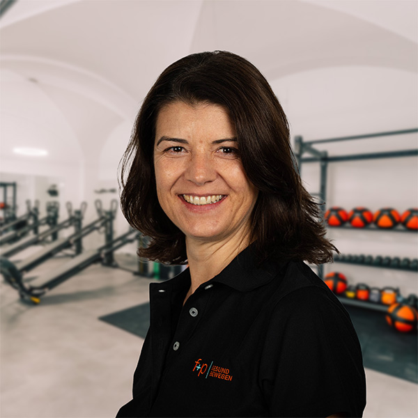 Frau mit dunklen Haaren in einem schwarzen Poloshirt lächelt vor Fitnessgeräten in einem Trainingsraum.