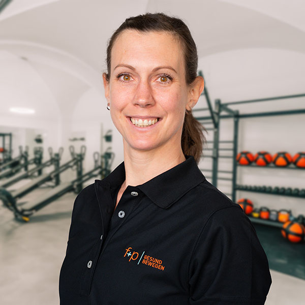 Frau mit braunen Haaren in schwarzem Polo-Shirt mit Logo von f+p Gesund Bewegen, lächelnd vor einem Fitnessbereich.