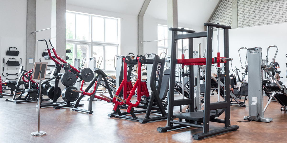 Fitnessstudio mit verschiedenen Trainingsgeräten, darunter Kraftmaschinen und Hanteln in einem hellen Raum.
