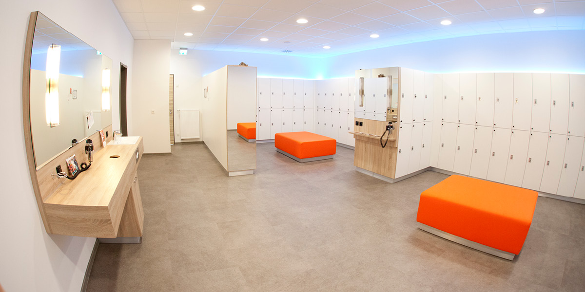 Umkleideraum mit orangefarbenem Sitzbereich und weißen Spinden in einem modernen Fitnessstudio.