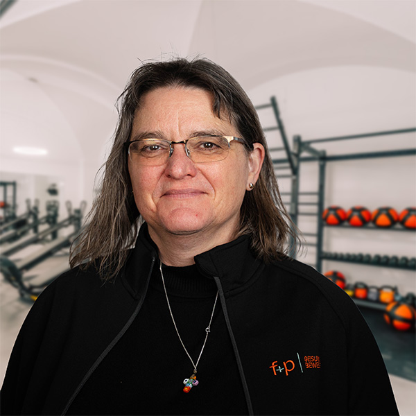 Frau mit Brille und schwarzem Oberteil, lächelnd vor einer Fitnessumgebung mit Geräten im Hintergrund.