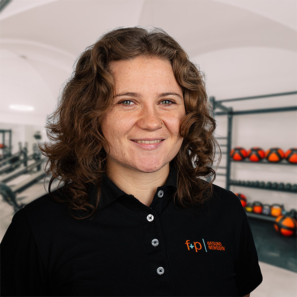 Frau mit lockigem Haar in schwarzem Poloshirt mit Logo von f+p Gesund Bewegen, lächelnd vor einem Fitnessraum.