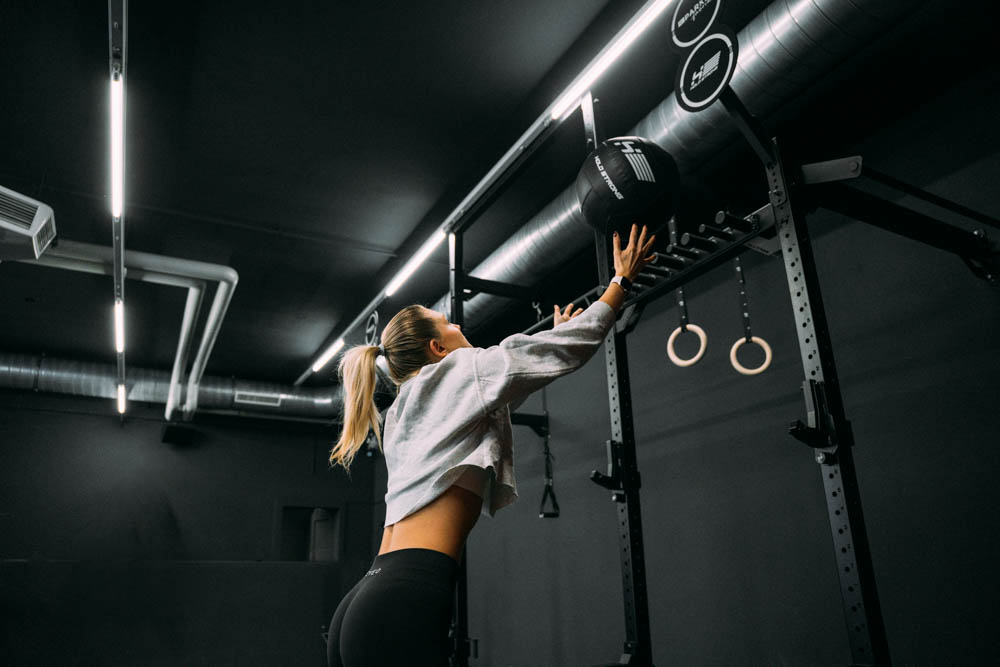 Frau hebt einen Medizinball an einer Trainingsstation in einem modernen Fitnessstudio mit dunkler Wandfarbe.