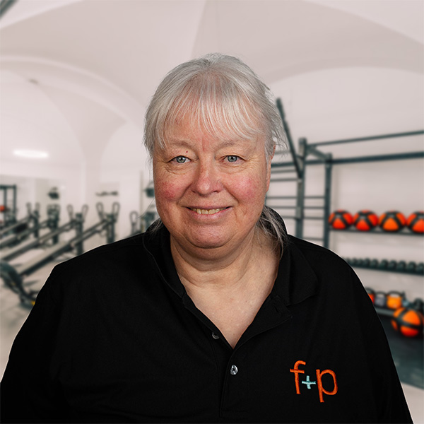 Frau mit grauen Haaren in schwarzem Polo-Shirt mit f+p Logo, lächelt vor einem Fitnessraum mit Geräten.