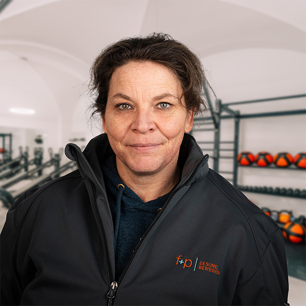 Frau mit dunkler Jacke und lässigem Pullover, die in einem Fitnessraum steht, im Hintergrund Gewichte sichtbar.
