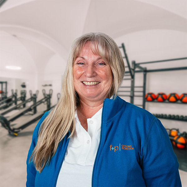 BriBu in blauer Jacke mit Logo von f+p Gesund Bewegen lächelt in einem Fitnessraum mit Geräten im Hintergrund.