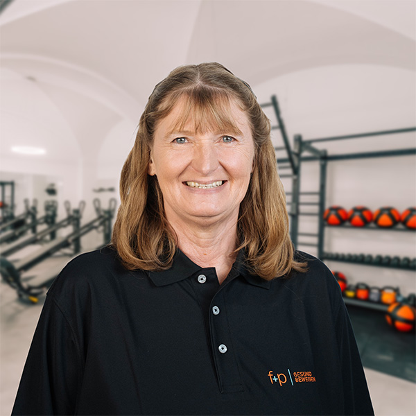 Frau mit langen, braunen Haaren in einem schwarzen Poloshirt vor einem Fitnessstudio mit Trainingsgeräten.