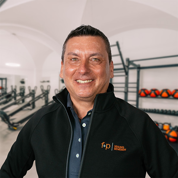 Mann in schwarzer Jacke mit Logo von f+p Gesund Bewegen, lächelnd in einem Fitnessraum.