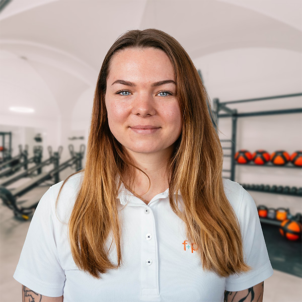 Frau mit langen, braunen Haaren in einem weißen Poloshirt von f+p Gesund Bewegen, lächelt in einem Fitnessstudio.