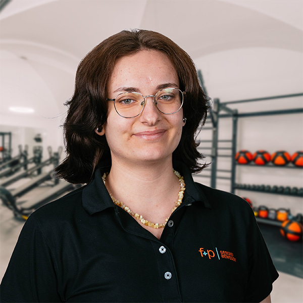 Frau mit Brille und schwarzem Polohemd mit f+p Gesund Bewegen Logo in einem Fitnessstudio mit Gewichten im Hintergrund.