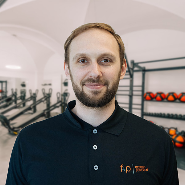Porträt von David, Mitarbeiter bei f+p Gesund Bewegen im Allgäu, mit dunkelblauem Polo und Bart, vor Trainingsgeräten im Fitnessraum.