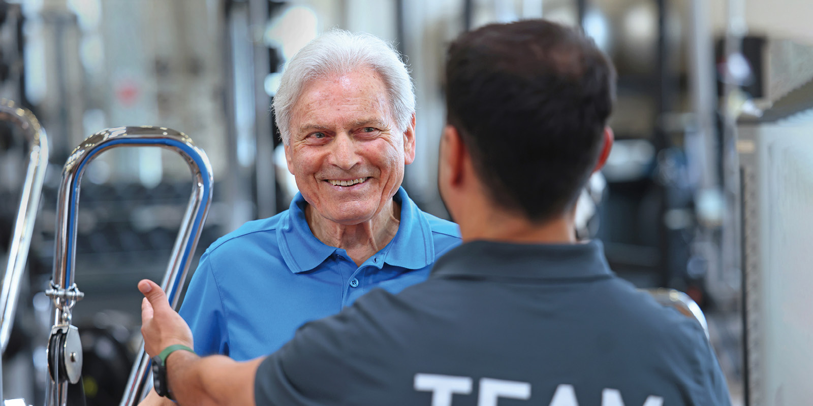 Ein älterer Mann im Fitnessstudio im blauen Polohemd im Gespräch mit Trainer an Kraft- und Cardio-Geräten, Fokus auf Training bei f+p Gesund Bewegen.