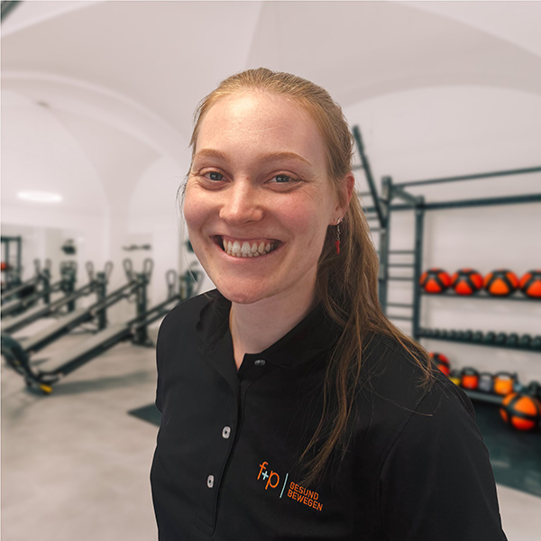 Johanna lächelt in einem schwarzen Poloshirt von f+p Gesund Bewegen in einem modern ausgestatteten Fitnessbereich.