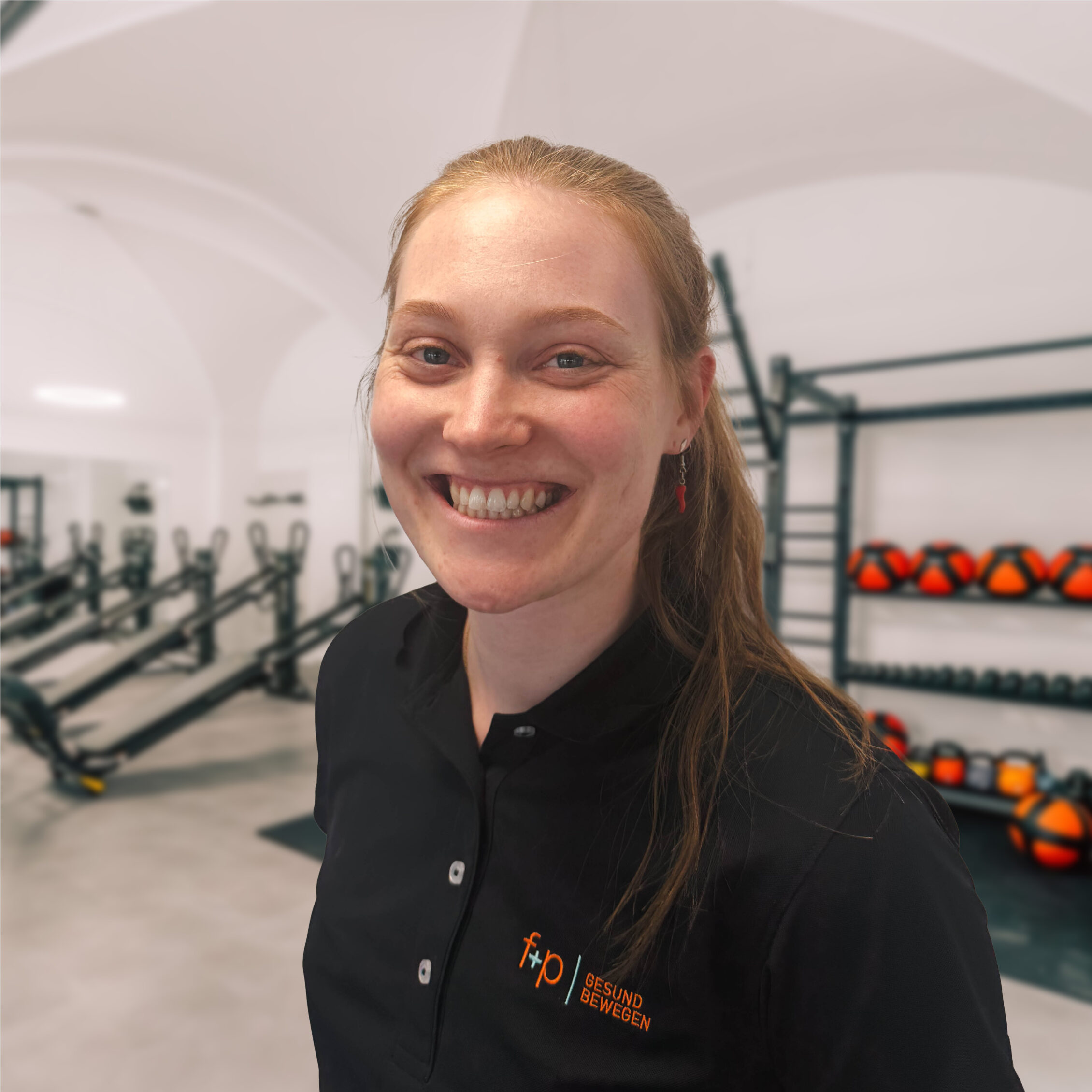 Lächelnde Johanna von f+p Gesund Bewegen im Bereich Physiotherapie, mit blondem Haar und dunkler Arbeitskleidung in der Trainingshalle.