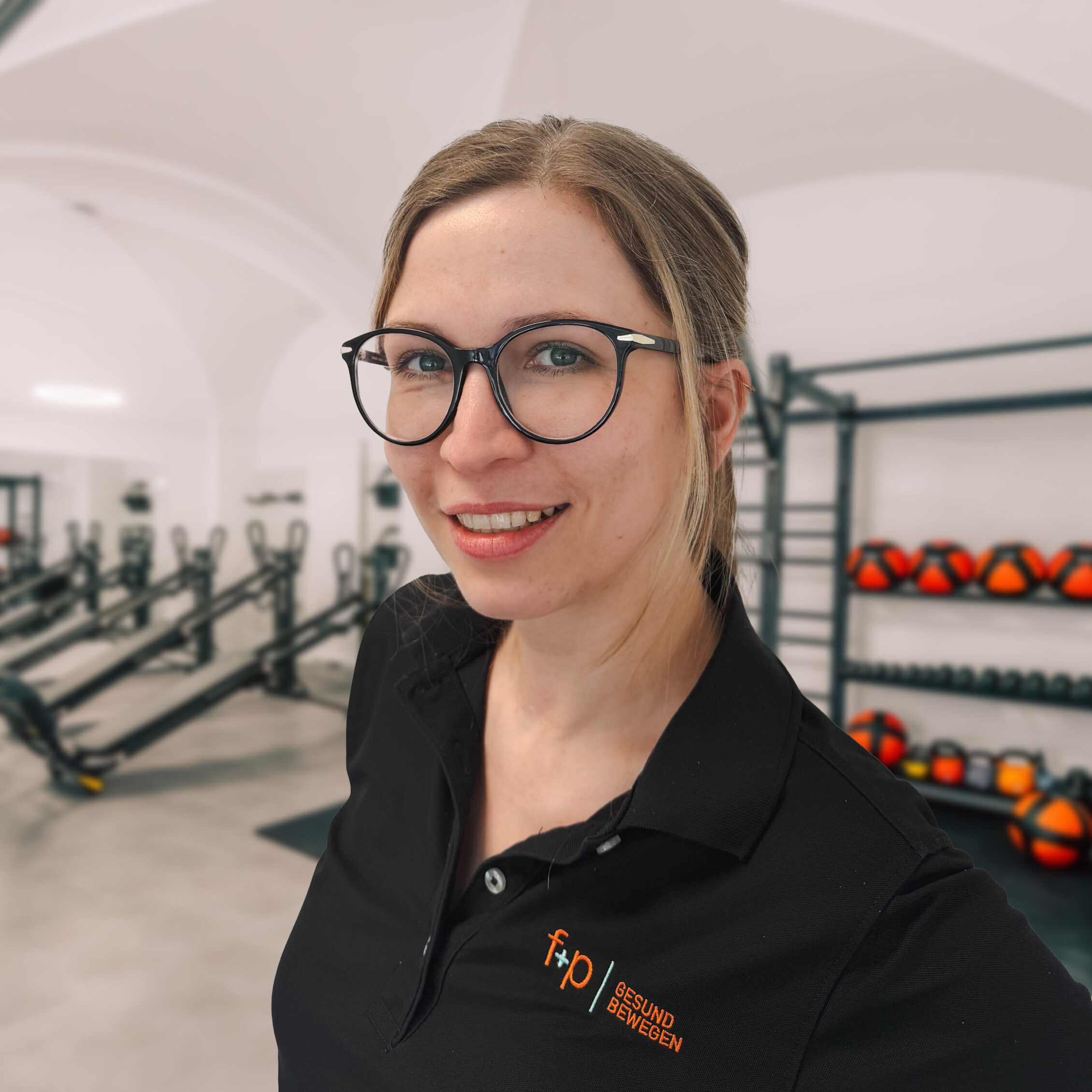 Julia mit schwarzer Brille und blondem Haar in der Praxis von f+p Gesund Bewegen, im Hintergrund Geräte und Trainingstools.
