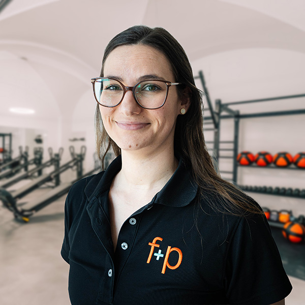 Marie-Theres von f+p Gesund Bewegen trägt eine Brille und ein schwarzes Polo mit f+p Logo im Trainingsraum für Physiotherapie im Allgäu mit Sportgeräten.