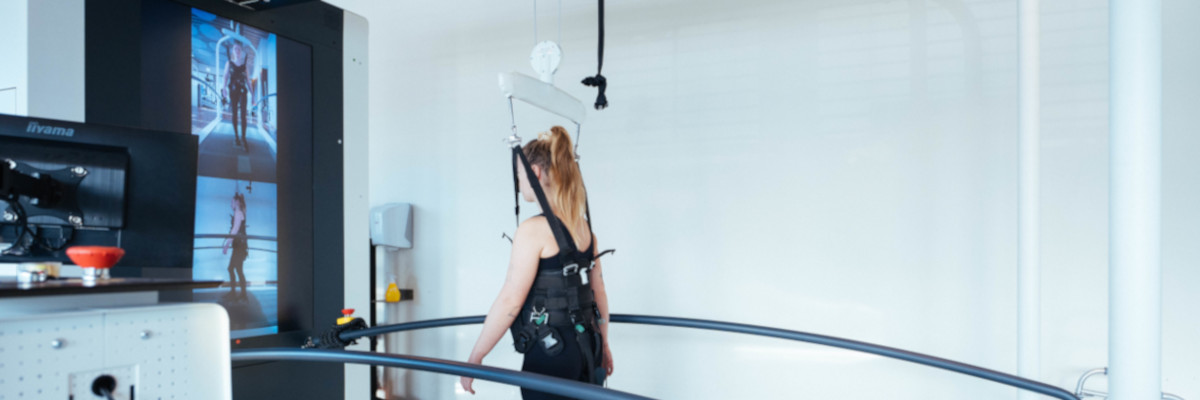 Im Entlastung C-Mill Reha-System geht eine Person mit Stützgurt auf einer Laufbahn; Monitorbilder zeigen Daten zum Gangbild.