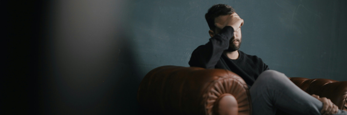 Psychosomatische Reha: Eine gestresste Person in dunkler Kleidung hält sich die Stirn im Gespräch, blaugrauer Hintergrund und angespannter Gesichtsausdruck.