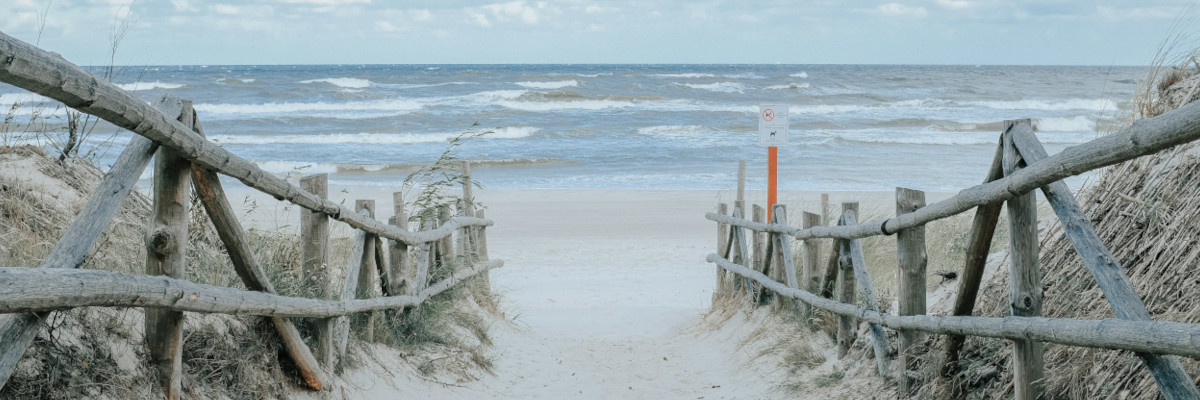 Blick durch einen Holzschutzzaun auf sandige Dünen und die raue Nordsee mit auflaufenden Wellen unter wolkigem Himmel.