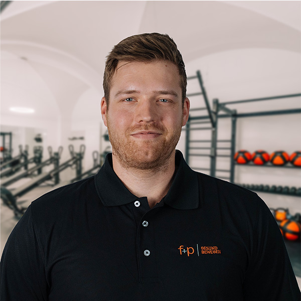 BaFl Bastian im dunkelblauen f+p Gesund Bewegen Poloshirt vor Trainingsgeräten im Allgäu, mit klar erkennbarer Team-Logoplatzierung.