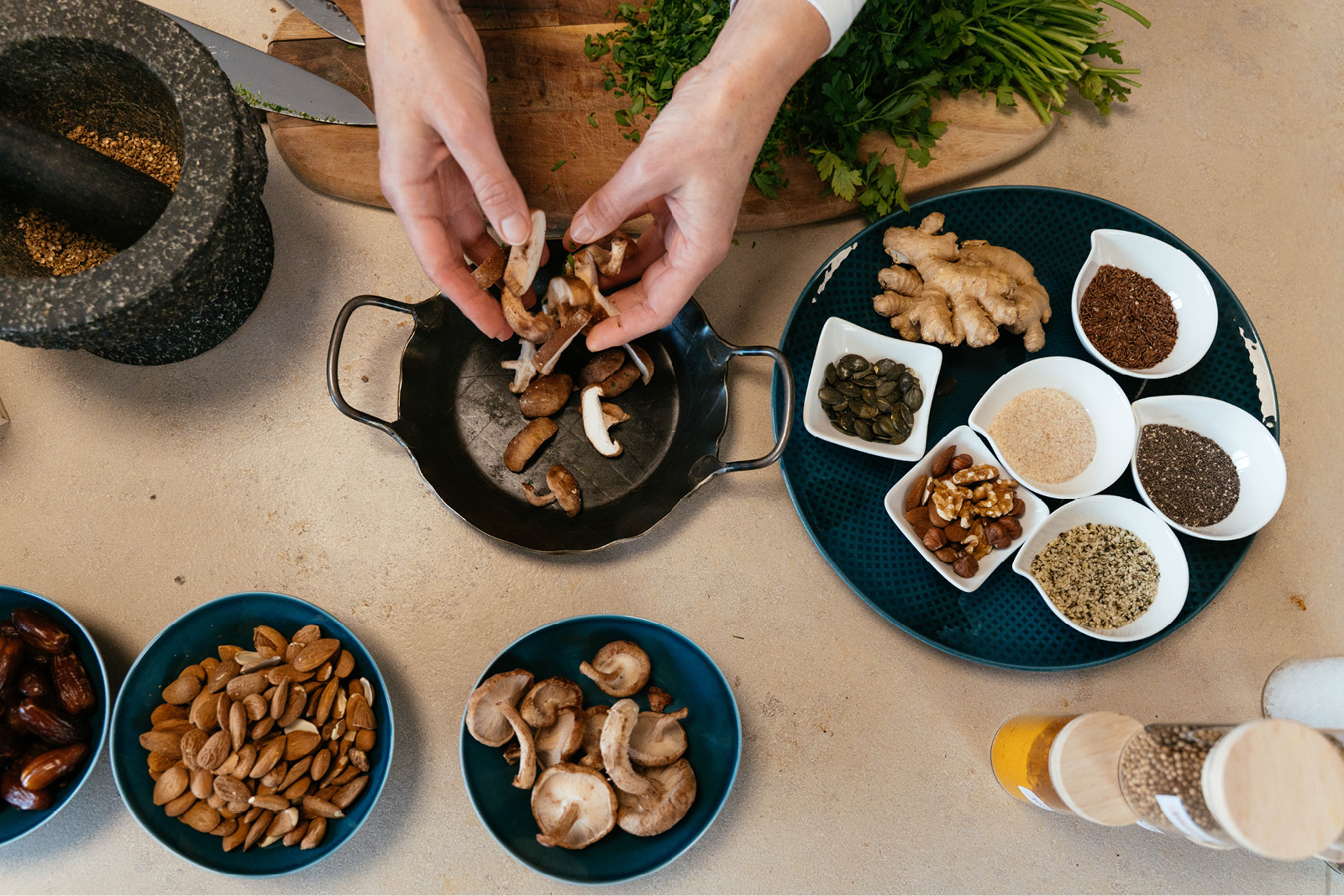Kochkurse-website Beim Kochen werden Champignons vorbereitet: Hände schneiden Pilze auf dem Brett, dazu stehen Gewürzschalen mit grünen Kräutern und Samen bereit.