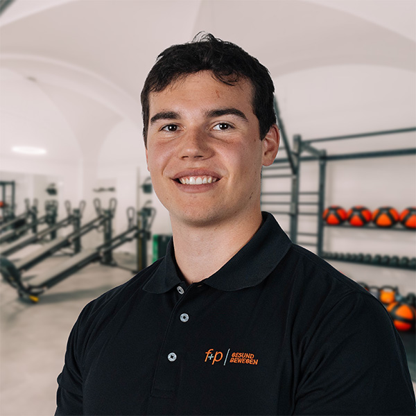 Moritz von f+p Gesund Bewegen im schwarzen Poloshirt mit freundlichem Lächeln im Reha-Training-Bereich.