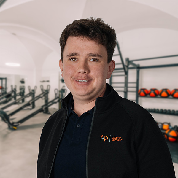 MoGr Moritz mit schwarzer Jacke und f+p Gesund Bewegen Logo im Therapie- und Reha-Bereich, kurzhaarig, im Hintergrund Geräte.