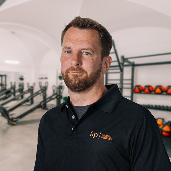 Moritz mit Bart und schwarzem Poloshirt im Trainingsraum von f+p Gesund Bewegen, mit orangefarbenem Logo.