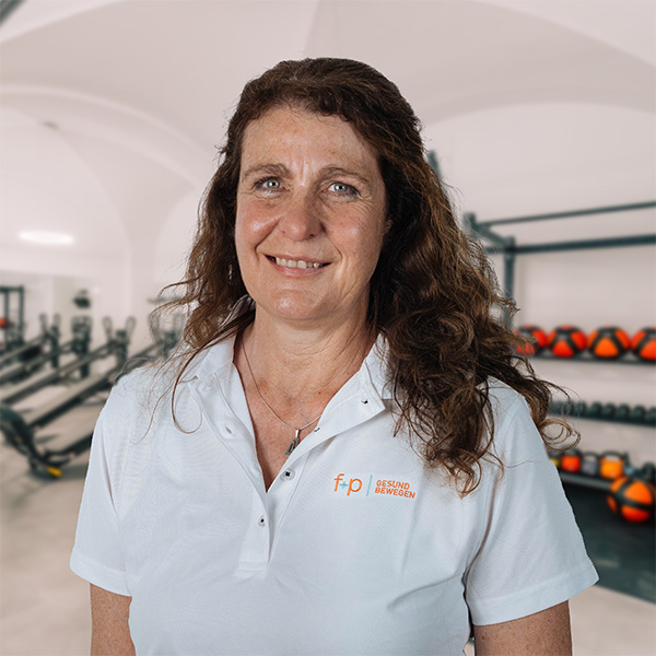 Lächelnde Sabine von f+p Gesund Bewegen im Allgäu im weißen Shirt mit Logo, im Hintergrund Trainingsgeräte und ein heller Raum