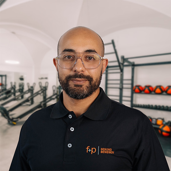 Im f+p Gesund Bewegen in Kempten steht Chakib vor Trainingsgeräten und Hanteln und trägt ein schwarzes Polo mit dem Logo von f+p Gesund Bewegen.
