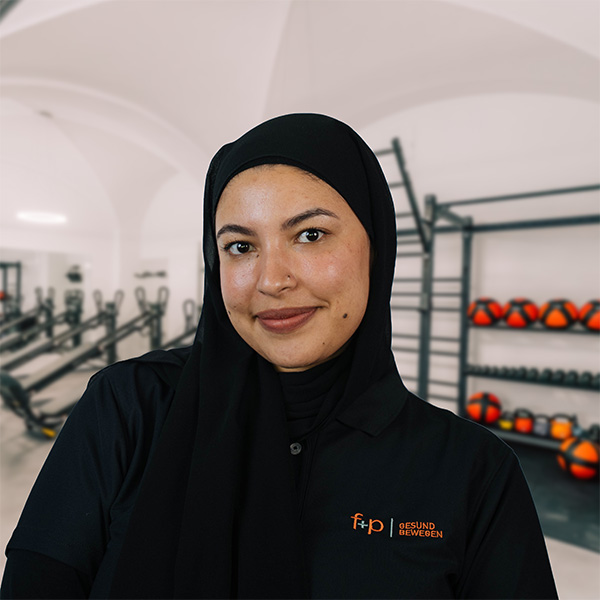 In der Physiotherapie bei f+p Gesund Bewegen steht Nour mit schwarzem Hijab und schwarzem Polo vor modernen Trainingsgeräten im Kemptener Studio und lächelt.