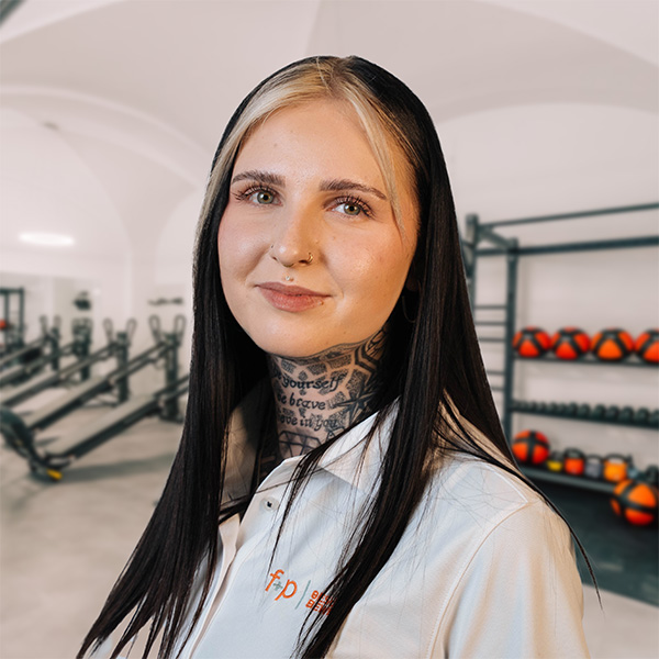 In der f+p Gesundheitsberatung steht Sophia im Trainingsraum vor Geräten und Medizinbällen, mit langen schwarzen Haaren und weißem Oberteil.