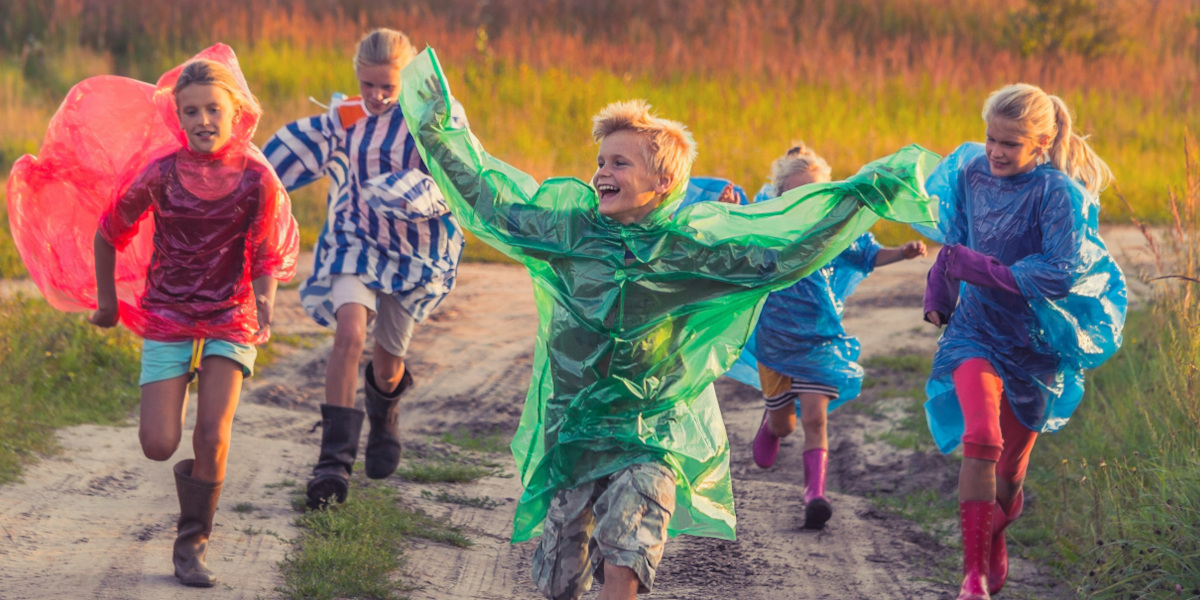 Mehrere Kinder rennen lachend über einen Feldweg und tragen bunte Regenponchos in Rot, Grün und Blau im warmen Abendlicht.
