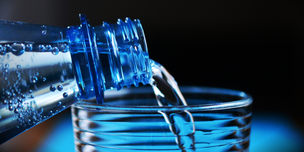 Clear water pours from a blue plastic bottle into a glass against a dark background, creating a cool, refreshing scene with bright reflections.