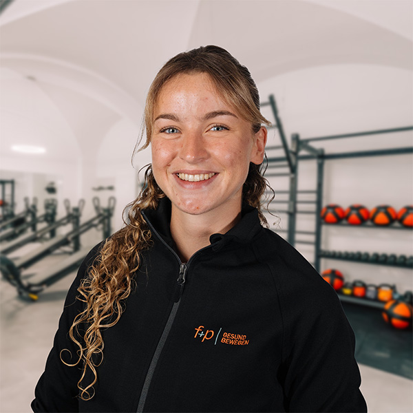 Eine freundlich lächelnde Therapeutin von f+p Gesund Bewegen steht vor Trainingsgeräten in einer hellen Praxis mit orangefarbenen Bällen.
