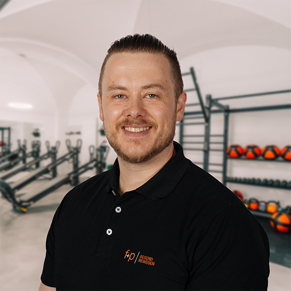 Michael von f+p Gesund Bewegen lächelt in schwarzem Polo in einem hellen Trainingsraum mit Kraftgeräten, Racks und Kettlebells im Hintergrund.
