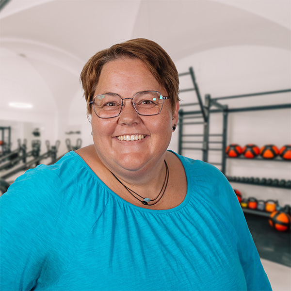 Lächelnde Samantha mit Brille und blauem Oberteil steht in einem hellen Trainingsraum mit Therapiegeräten und Medizinbällen im Hintergrund.