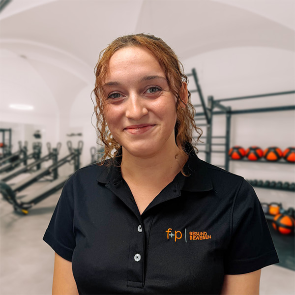 Eine Mitarbeiterin von f+p Gesund Bewegen lächelt in einem schwarzen Poloshirt im Trainingsbereich mit Fitnessgeräten.
