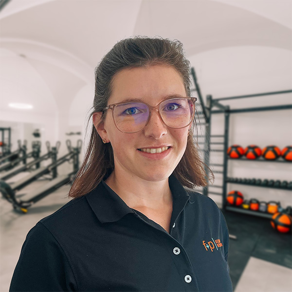 Claudia lächelt in schwarzem Poloshirt mit f+p-Logo in einem Physiotherapieraum mit Trainingsgeräten und orangefarbenen Bällen.