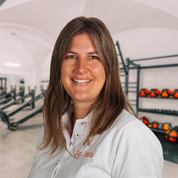 LieHa Lächelnde Liesa von f+p Gesund Bewegen im weißen Hemd vor Geräten und Trainingsbällen in einem Fitnessbereich