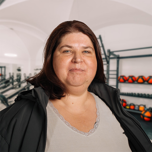Porträt von Luigia mit braunen Haaren und schwarzem Mantel in einem Fitnessbereich von f+p Gesund Bewegen