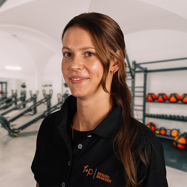 Nadine mit braunem Haar und schwarzem f+p-Poloshirt in einem modernen Fitness- und Therapiebereich von f+p Gesund Bewegen.