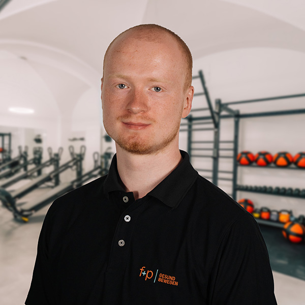PeWo Peter trägt ein schwarzes Poloshirt mit f+p-Logo und steht lächelnd im Trainingsbereich der neurologischen Therapie.