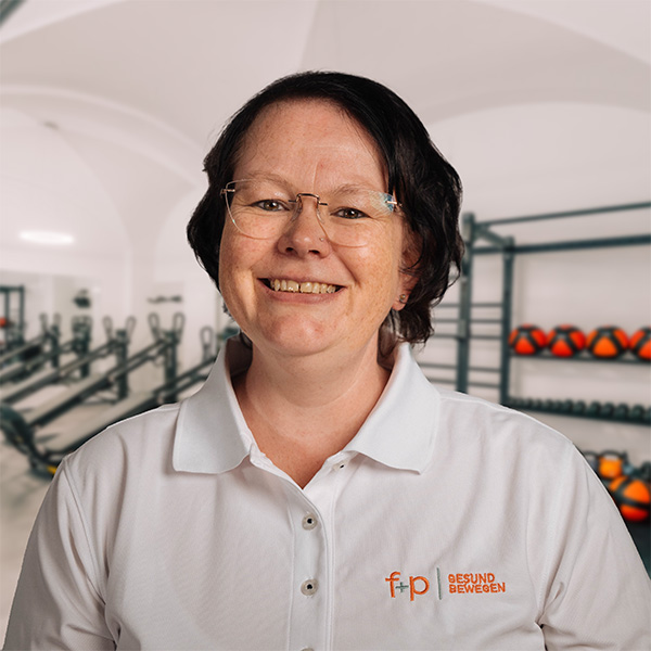 Freundlich lächelnde Yvonne im weißen Poloshirt mit f+p-Logo vor Geräten eines modernen Fitnessbereichs in Kempten.