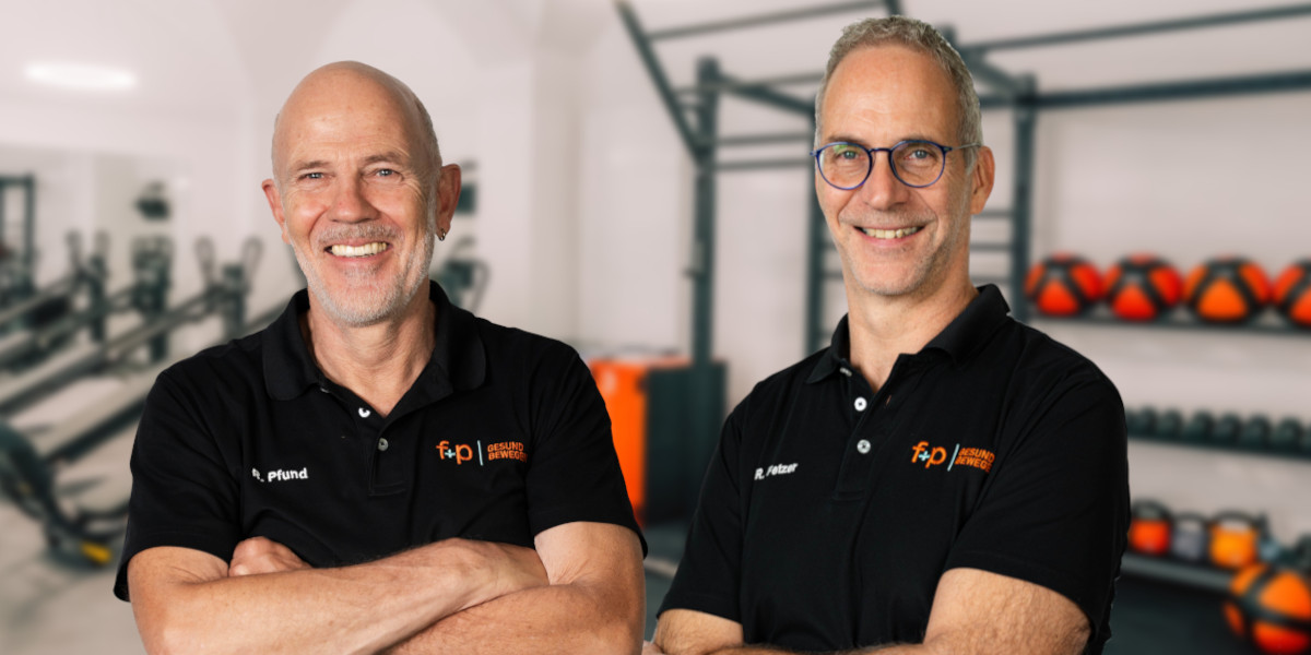 Zwei lächelnde Mitarbeiter von f+p Gesund Bewegen stehen in schwarzer Poloshirt-Kleidung in einem Physiotherapie-Trainingsraum.
