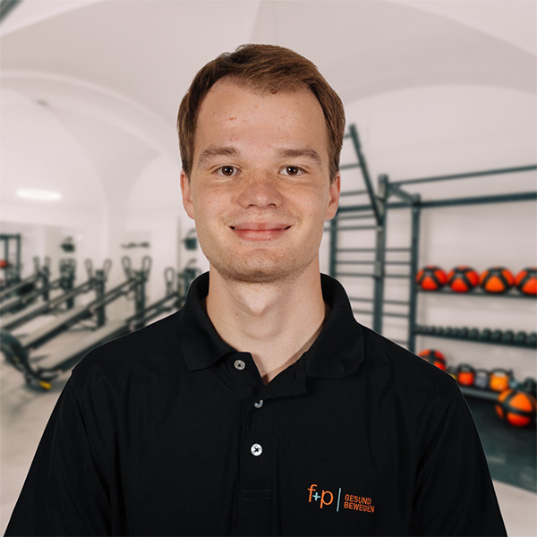 Ein lächelnder Mitarbeiter von f+p Gesund Bewegen steht im schwarzen Poloshirt in einem Trainingsraum mit Fitnessgeräten.