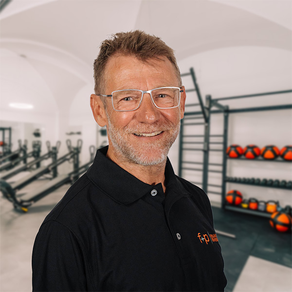 FrMi Ein lächelnder Mann mit rechteckiger Brille und schwarzem Poloshirt steht in einem Trainingsraum von f+p Gesund Bewegen.