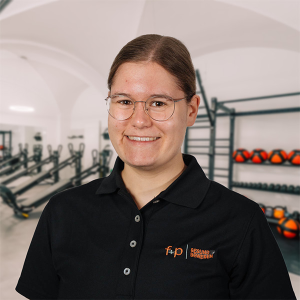 Leonie lächelt in schwarzem Poloshirt mit f+p-Logo in einem Trainingsbereich mit Fitnessgeräten und Ergotherapie-Ausstattung.